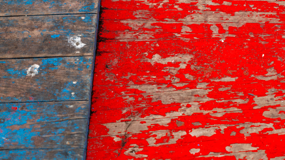Wooden planks in blue and red which are heavily weathered. The colour peels off strongly. The wood can be seen under the paint.