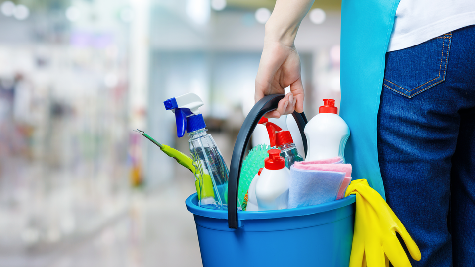 A cleaning woman is standing inside a building holding a blue bucket filled with chemicals and facilities for tidying up in her hand.